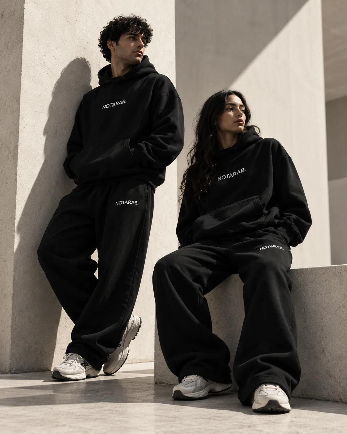 Two people wearing NOTARAB. black hoodies and sweatpants against a sunlit concrete wall